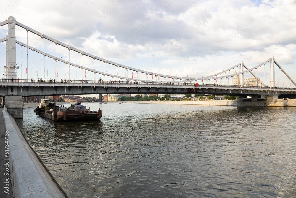 Moscow river with barge and cargo, pedestrian bridge with pedestrians. Summer day, cloudy sky. Center of Moscow. Moscow river with barge and cargo, pedestrian bridge with pedestrians. Summer day, cloudy sky. Center of Moscow.