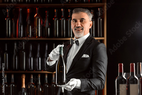Elegant waiter sommelier wine steward with a bottle of wine. Examination of wine products. Restaurant staff among shelves of wine bottles. Stylish middle-aged man with a grey beard.