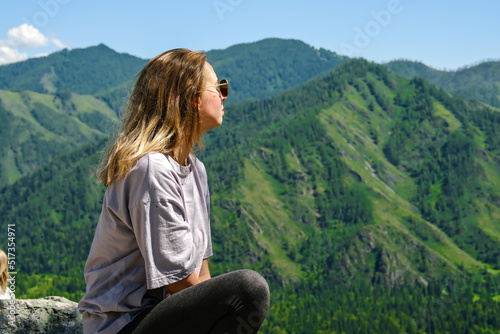 A young girl enjoys the nature of the mountains. Landscapes of mountains. A girl with blonde hair sits and looks at the mountains.