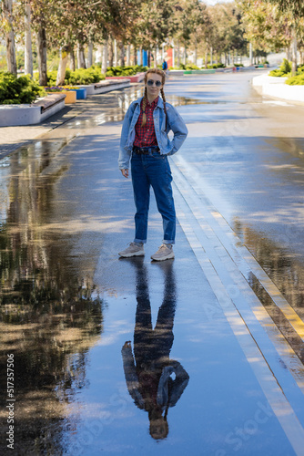A woman with pigtails in a youth denim outfit is reflected in a puddle of wet avfalt in a spring park. Walk after the rain.