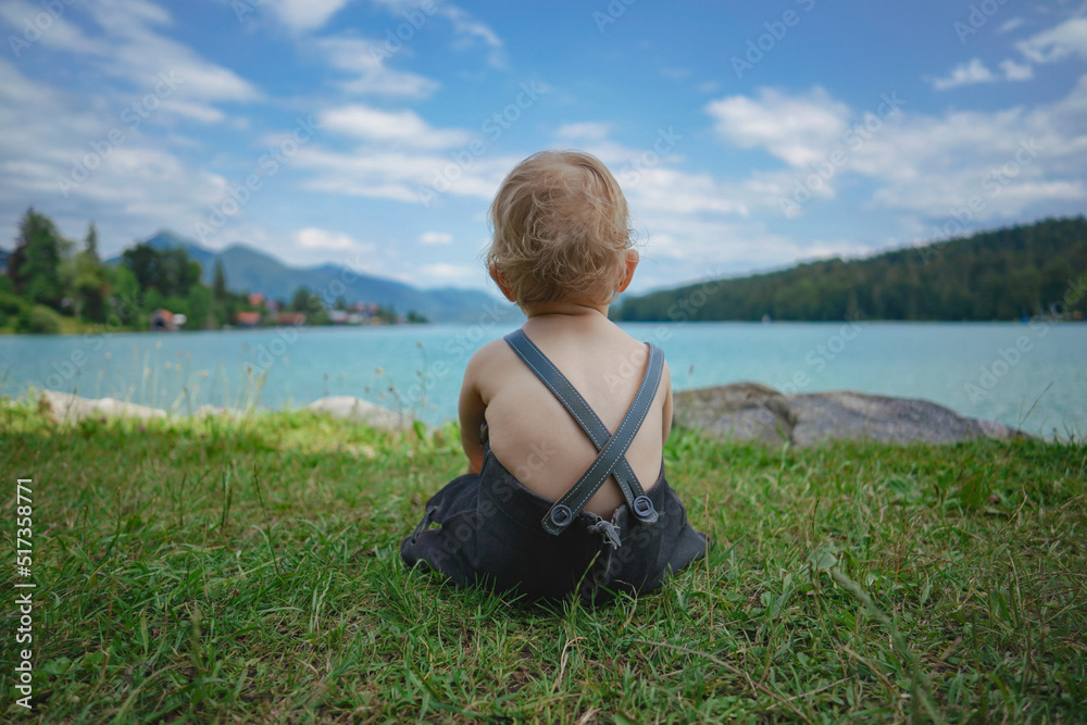 back of a blond german baby boy in bavarian dress with lederhose sitting on the meadow by the shore of the famous bavarian lake Walchensee with turquoise blue water in the background and watching 