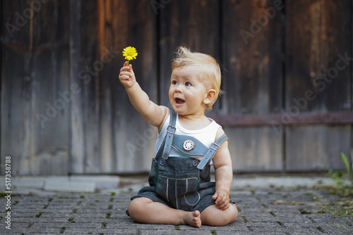 HAPPY SMILING BABY OF 1 YEAR SITTING  IN BAVARIAN LEATHER PANTS called Lederhosn OUTDOORS ON THE FLOOR WITH ARMS RAISED AND HOLDING A FLOWER IN THE AIR TO CONGRATULATE photo