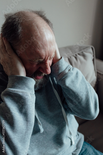 Ukrainian worried middle aged man was frightened by the loud sound and closed his ears in fear. Unhappy mature older grandfather at home.