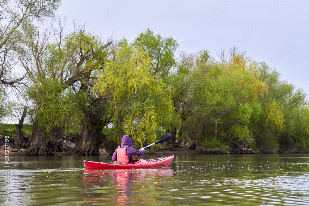 Woman in red kayak paddle at river near shoreline with green gentle spring trees