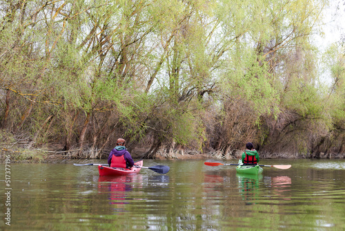 Rear view of two women sitting in red and green kayaks. Spring Danube river near trees with gentle green leaves