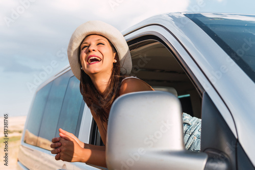 Young girl with hat leaning out the window of her camper van and smiling energetically. Brunette woman looking at the scenery of a countryside area on a summer road trip. Concept of van life.