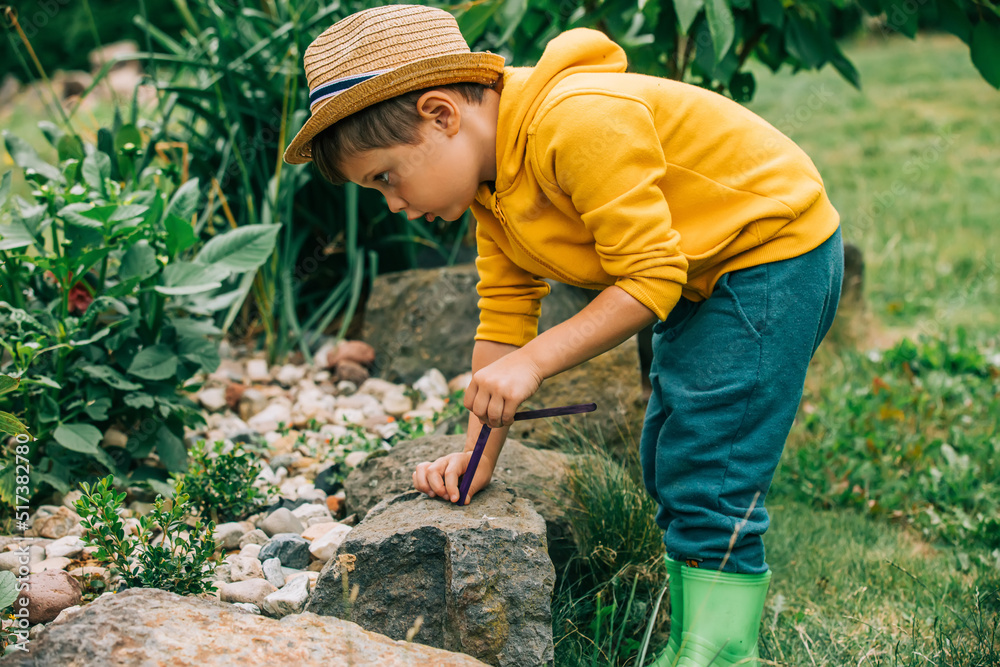 Fototapeta premium Little boy in hat and yellow jacket plays in a garden