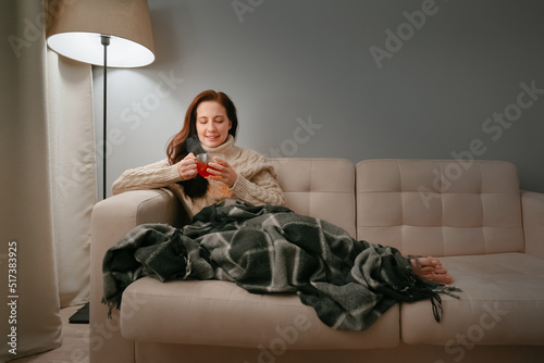 Vászonkép Young woman wrapped herself in a plaid while sitting on a beige sofa drinks hot tea for colds
