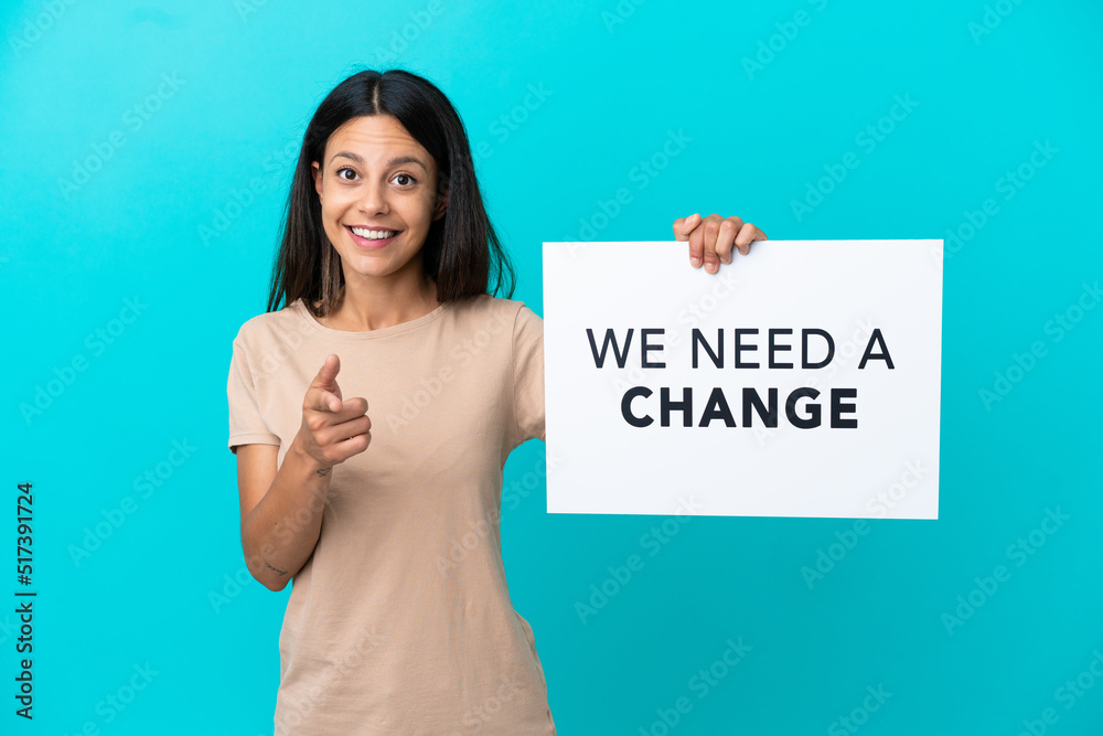 Young woman over isolated background holding a placard with text We Need a Change and pointing to the front Young woman over isolated background holding a placard with text We Need a Change and pointing to the front