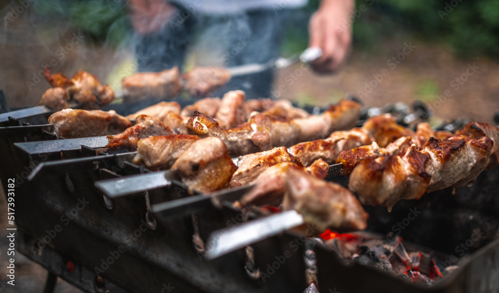 Man hand straightens the skewer with barbecue delicious grilled meat on brazier outdoors. Person preparing pork steaks with smoke during picnic at the nature. Hot BBQ closeup