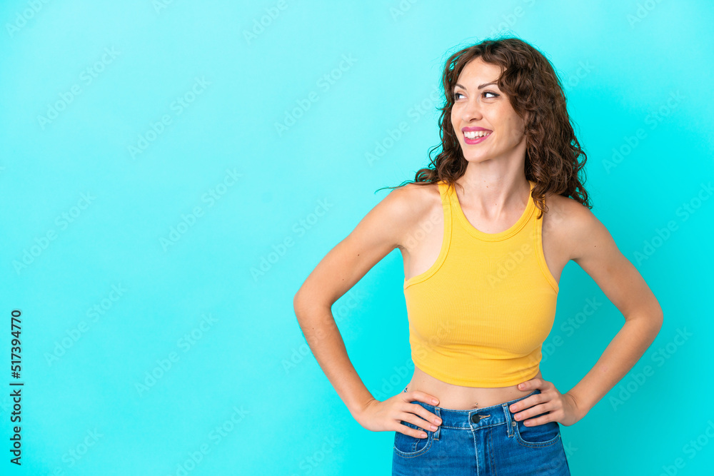 Young woman with curly hair isolated on blue background posing with arms at hip and smiling Young woman with curly hair isolated on blue background posing with arms at hip and smiling