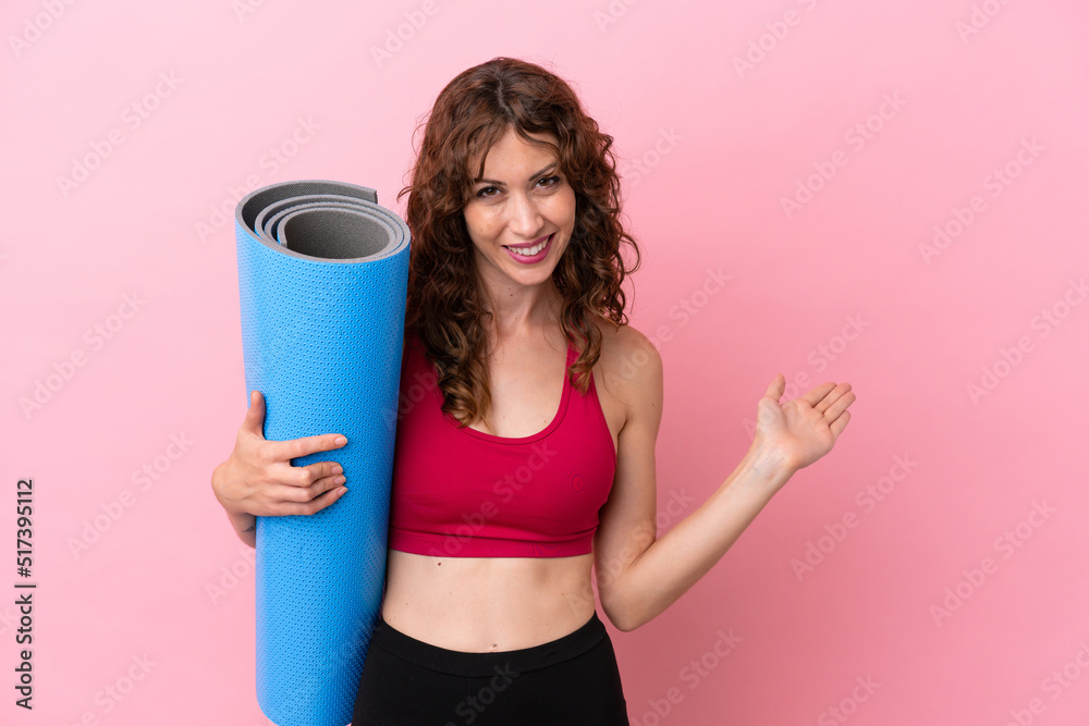 Young sport woman going to yoga classes while holding a mat isolated on pink background extending hands to the side for inviting to come Young sport woman going to yoga classes while holding a mat isolated on pink background extending hands to the side for inviting to come