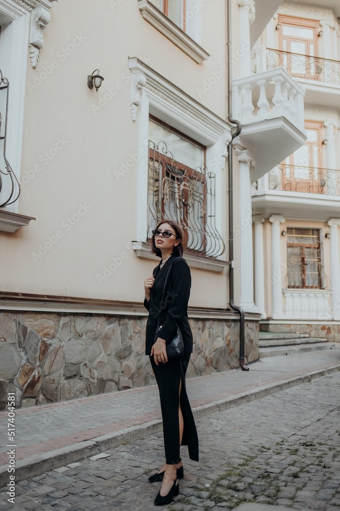 a brunette girl in a black jacket and dress and black glasses walks down the street in the city with a handbag a brunette girl in a black jacket and dress and black glasses walks down the street in the city with a handbag