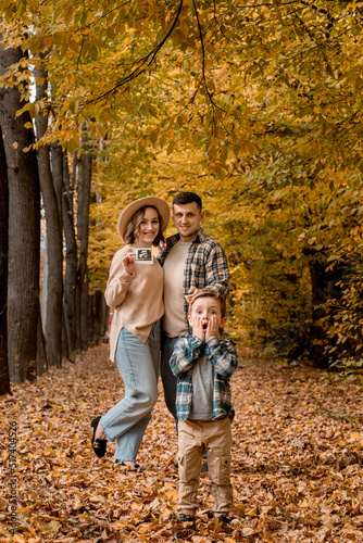 Happy family of a man with a woman holding an ultrasound pregnancy picture and a surprised son who will be the older brother. Autumn photo shoot of a family waiting to be replenished