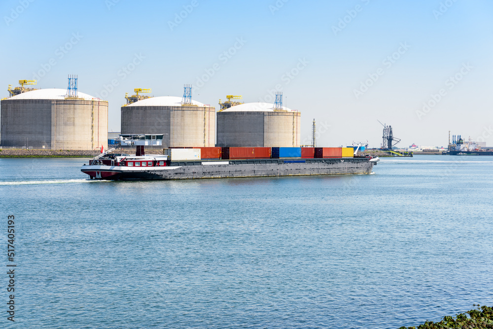 Container barge with harbourside lquefied natural gas tanks in background on a clear summer day. Port of Rotterdam Port, Netherlands. Container barge with harbourside lquefied natural gas tanks in background on a clear summer day. Port of Rotterdam Port, Netherlands.