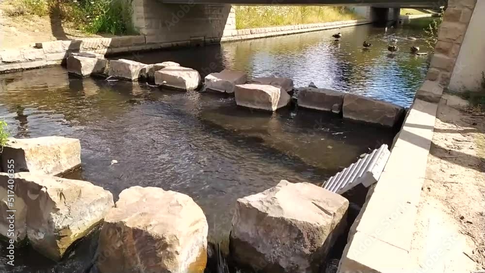 Little city creek with calm flowing water in slow motion through rocks in water under little bridge in summer in european city with ducks and clear water for relaxed recreation in urban environment Little city creek with calm flowing water in slow motion through rocks in water under little bridge in summer in european city with ducks and clear water for relaxed recreation in urban environment