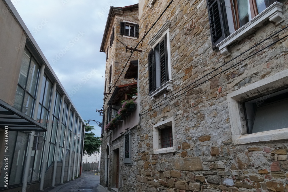 View on a narrow street with old stone building and new modern house in center of Koper, Slovenia. Between buildings is blue sky with white clouds. Old house has a balcony with flowers.