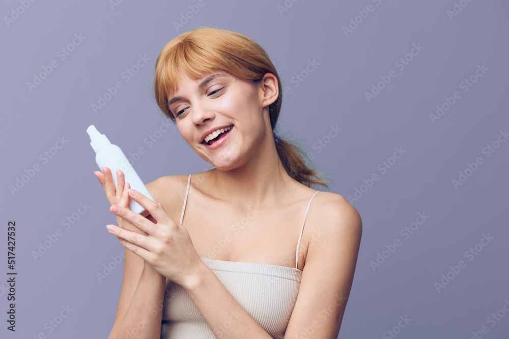 A joyful woman is surprised in a white T-shirt with fresh clean skin, with red wavy hair gathered in a tail, he stands on a lilac background with a white bottle in his hands.Horizontal studio shot. A joyful woman is surprised in a white T-shirt with fresh clean skin, with red wavy hair gathered in a tail, he stands on a lilac background with a white bottle in his hands.Horizontal studio shot.