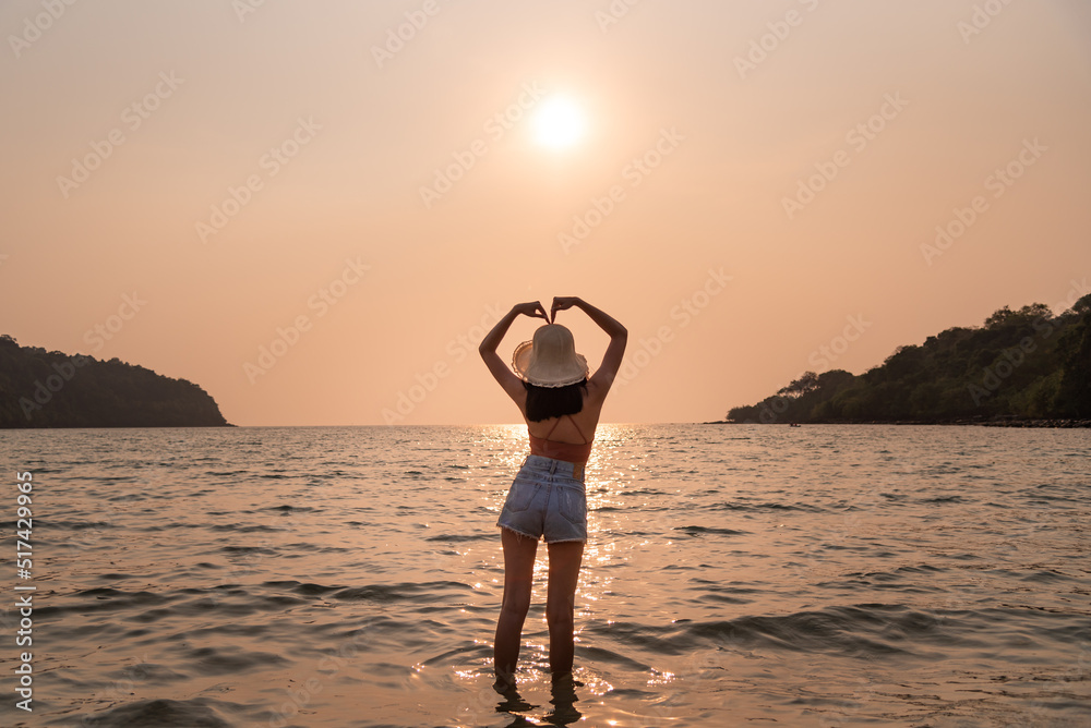 Alone girl show love symbol by hand at the sea among sunset