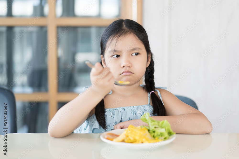 Asian child frowning or kid girl unhappy and don't like eating vegetables salad with corn tomato and greens vegetable with holding spoon or person anorexic bored food at kitchen school or restaurant Asian child frowning or kid girl unhappy and don't like eating vegetables salad with corn tomato and greens vegetable with holding spoon or person anorexic bored food at kitchen school or restaurant