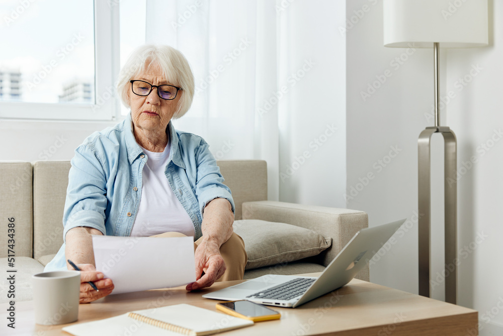an elderly lady works sitting on the couch at home reading a text from a paper during an online conference with colleagues an elderly lady works sitting on the couch at home reading a text from a paper during an online conference with colleagues