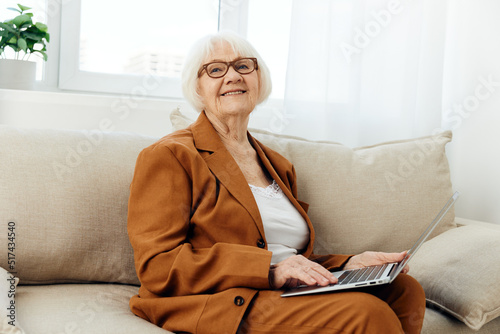 a funny elderly woman with gray hair is sitting in a bright room against the background of a window and biting her lower lip holds a laptop on her lap © Tatiana