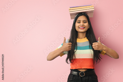 Excited smiling young indian asian  girl student posing islolated balancing books on her head and showing thumbs up with both hands pointing to a copyspace.