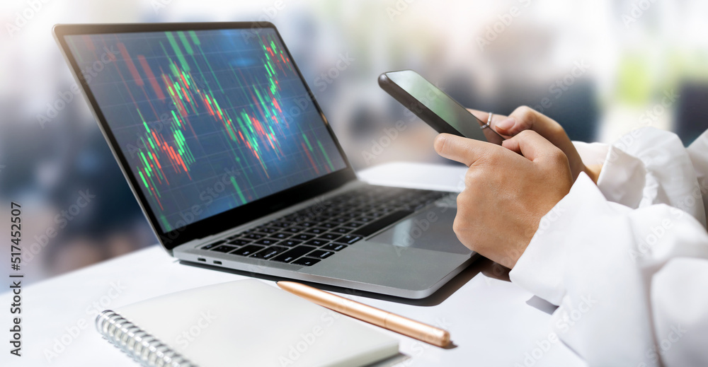 Young asian businesswoman using mobile phone to find information for investment analysis and analyze stock graph on laptop screen to make profitable plans. Close up view Young asian businesswoman using mobile phone to find information for investment analysis and analyze stock graph on laptop screen to make profitable plans. Close up view