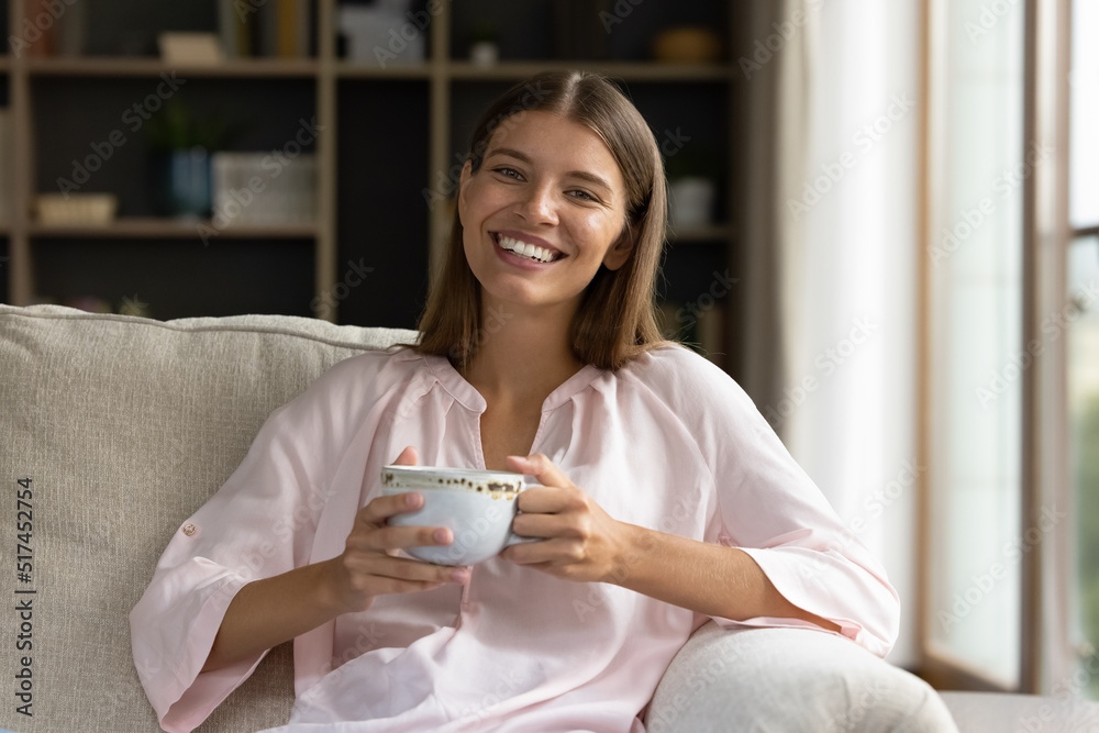 Young pretty woman resting on sofa in living room holds tea cup smile look at camera, enjoy morning coffee feel happy spend morning weekend leisure at cozy home, take break, relish pause from chores Young pretty woman resting on sofa in living room holds tea cup smile look at camera, enjoy morning coffee feel happy spend morning weekend leisure at cozy home, take break, relish pause from chores