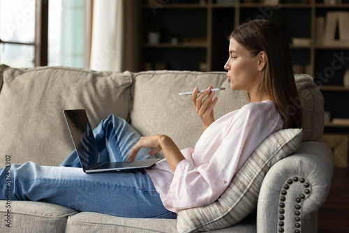 Side view woman holding cellphone makes personal or business call talks on speaker phone, web surfing information using laptop sit on sofa at home. Remote communication, young gen and AI tech concept