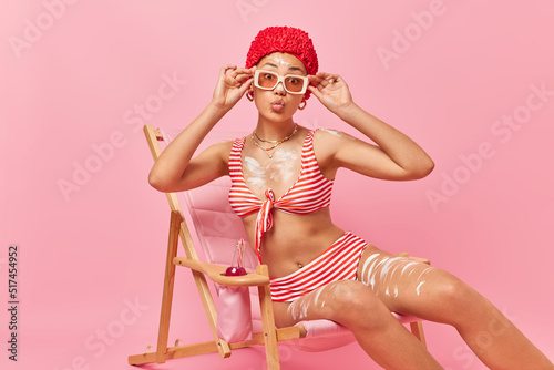 Lovely young woman applies sun protection cream wears hat striped swimsuit and sunglasses poses on comfortable deck chair enjoys summer vacation isolated over pink background. Holidays concept photo