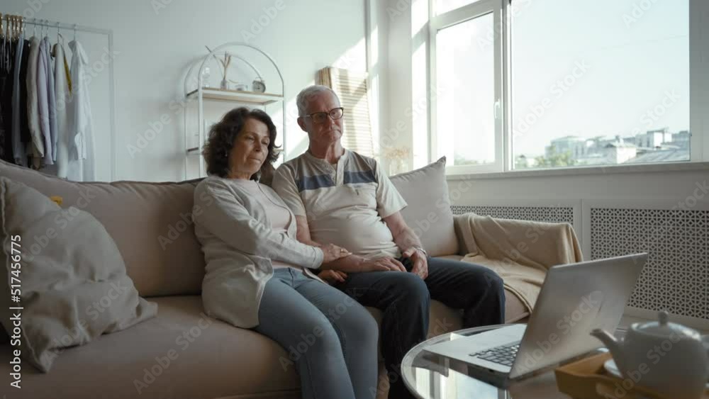 Elderly couple talks with children on video call in living room with big window. Senior man and woman hold hands sitting on comfortable sofa at home Elderly couple talks with children on video call in living room with big window. Senior man and woman hold hands sitting on comfortable sofa at home