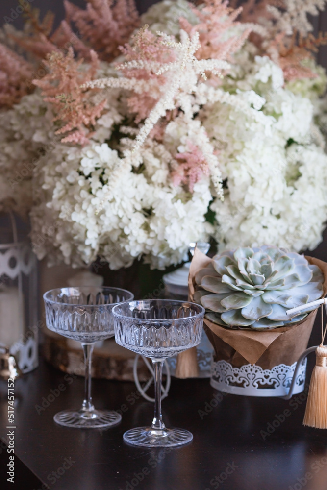 A bouquet of white hydrangeas and pink astilba, succulent, candles and a fashionable glass for dessert and champagne with wine inside. Cozy interior. The concept of a party, wedding and holiday. A bouquet of white hydrangeas and pink astilba, succulent, candles and a fashionable glass for dessert and champagne with wine inside. Cozy interior. The concept of a party, wedding and holiday.