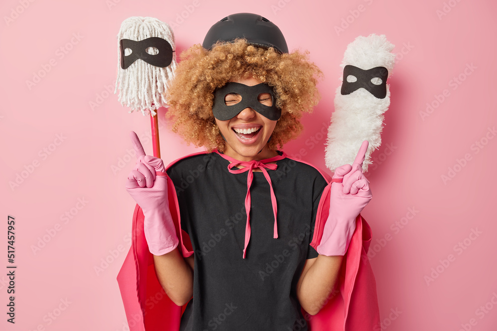 Cheerful curly woman happy to complete housework makes peace gesture smiles gladfully dressed in superhero costume poses with mop and brush behind isolated over pink background. Domestic chores Cheerful curly woman happy to complete housework makes peace gesture smiles gladfully dressed in superhero costume poses with mop and brush behind isolated over pink background. Domestic chores