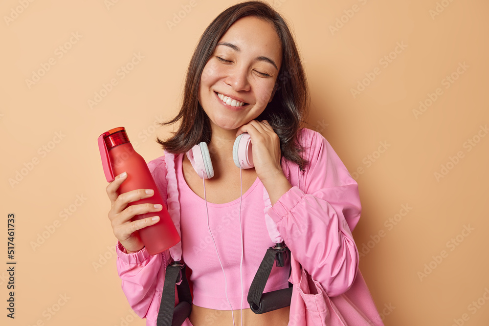 Φωτογραφία Horizontal shot of cheerful Asian woman dressed in sportswear holds bottle of water uses stereo headphones keeps eyes closed leads healthy lifestyle isolated over beige background