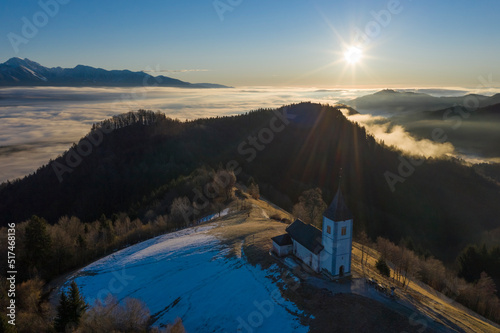 Church of St. Primoz and Felicijan at Sunrise above the Clouds. Julian Alps. Jamnik, Slovenia, Europe. Aerial View.