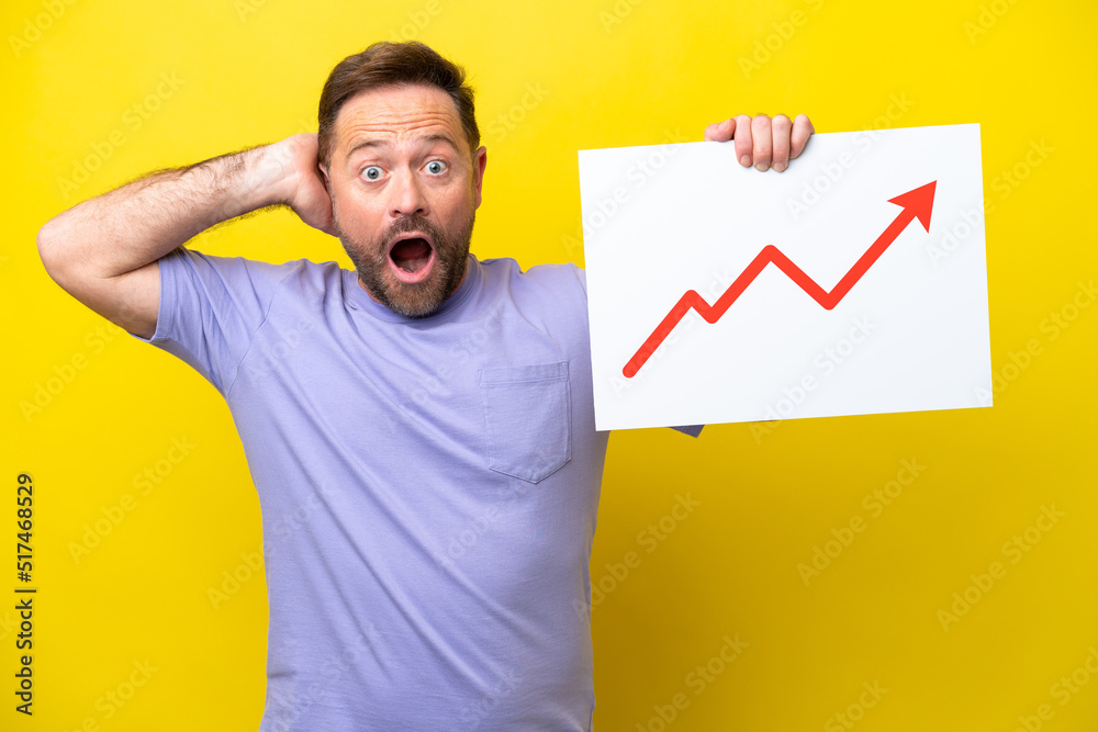 Middle age caucasian man isolated on yellow background holding a sign with a growing statistics arrow symbol with surprised expression Middle age caucasian man isolated on yellow background holding a sign with a growing statistics arrow symbol with surprised expression