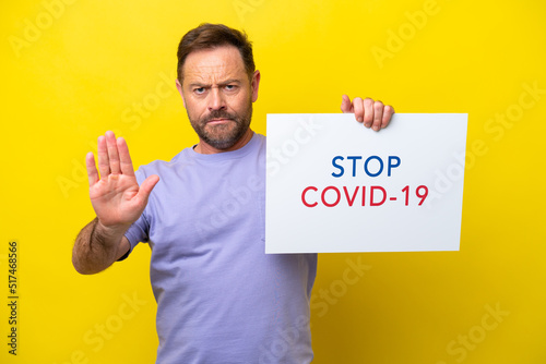 Middle age caucasian man isolated on yellow background holding a placard with text Stop Covid 19 and doing stop sign © luismolinero