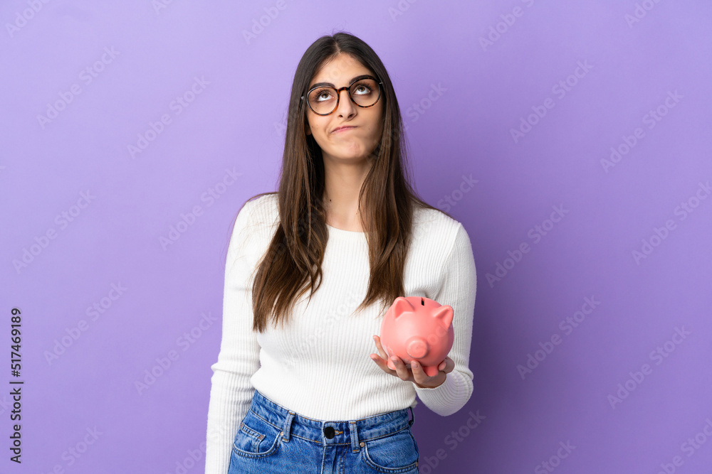 Young caucasian woman holding a piggybank isolated on purple background and looking up Young caucasian woman holding a piggybank isolated on purple background and looking up