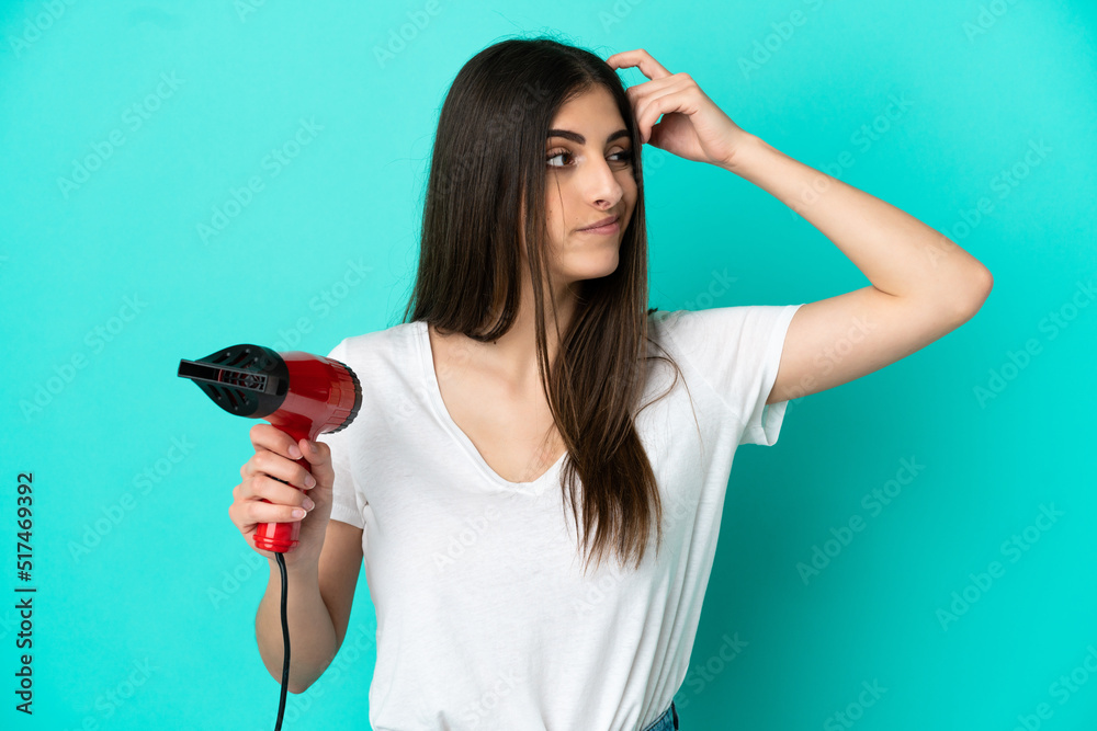 Young caucasian woman holding a hairdryer isolated on blue background having doubts and with confuse face expression Young caucasian woman holding a hairdryer isolated on blue background having doubts and with confuse face expression