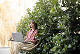 Young woman works on laptop computer while sitting relaxed on chair on background of green bushes at backyard. Concept of remote work at cozy atmosphere