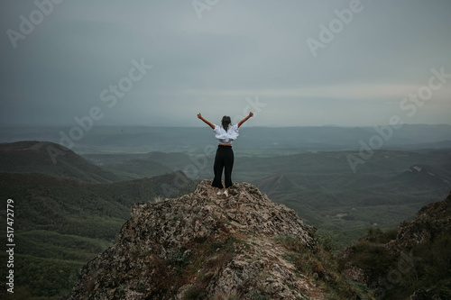 Happy woman tourist hiking up mountain enjoying nature. Landscape in cloudy gloomy windy weather. Female in white shirt with open arms outstretched in joy  enjoying travel. Freedom concept