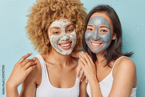 Cheerful women apply beauty clay mask for reducing blackheads smile gladfully stand closely to each other dressed in t shirt isolated over blue background. Skin care and wellness procedures.