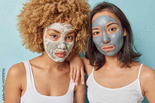 Horizontal shot of two mixed race young women apply skin care beauty mask dressed in t shirt stand closely to each other isolated over blue background. Women wellness and rejuvenation concept