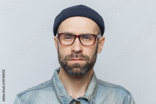 Canvastavla Photo of intelligent confident stylish man with dark thick beard and mustache, looks seriously into camera, poses against grey studio background, wears spectacles and hat