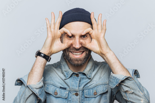 Portrait of hppy bearded male makes eyewear with fingers, has stubble, wears jeans cloting, stands against white background. Funny middle aged man foolishes indoor, pretends wearing glasses. photo