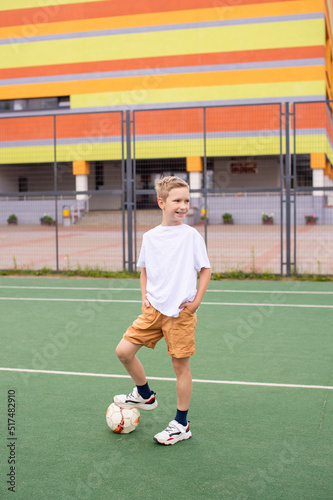 A teenage boy stands on a green field in the school yard with a soccer ball and looks away