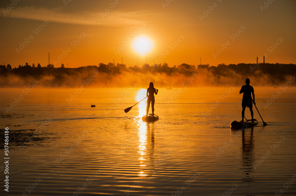 silhouettes of a couple on surfboards on the water at sunrise, fog on the horizon. SUP surfing at sunset