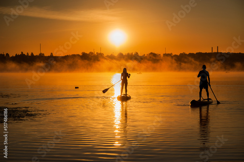 silhouettes of a couple on surfboards on the water at sunrise, fog on the horizon. SUP surfing at sunset