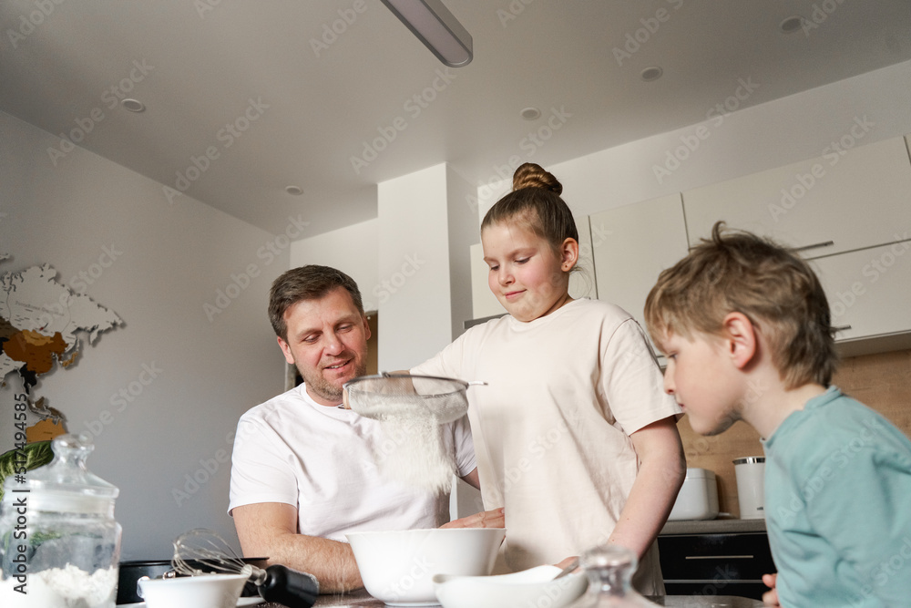 Obraz premium Dad and little daughter and son baking together in kitchen, preparing dough for pie, having fun at home during quarantine